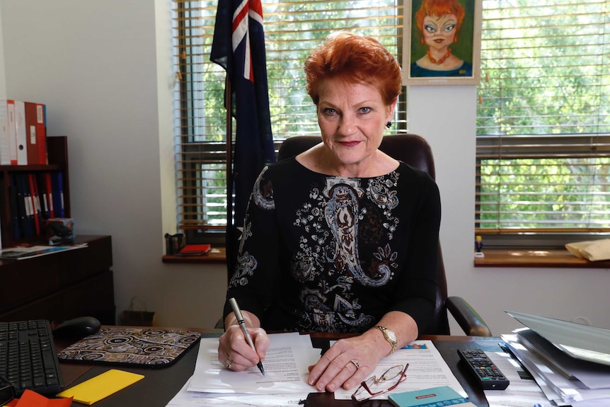 Middle aged woman sitting at desk with pen poised over papers.