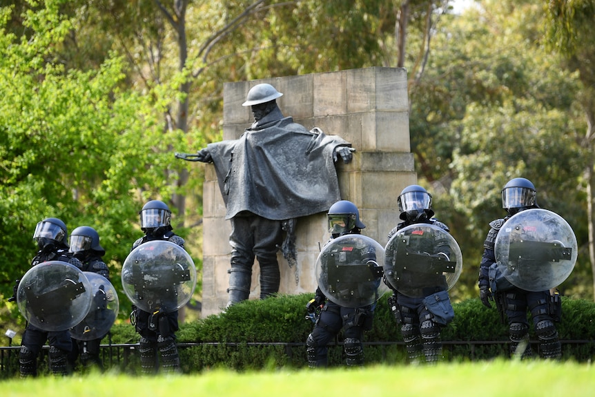 Riot police with shields and helmets stand in a line at the Shrine in front of a brass sculpture of a soldier.