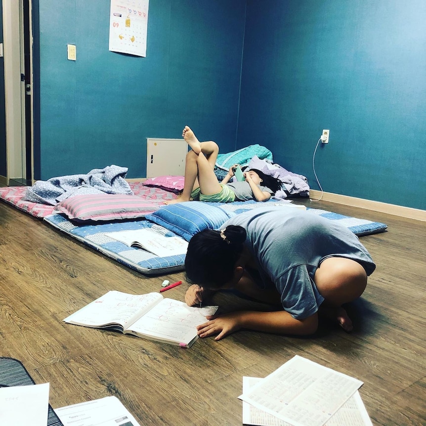 One young girl sits cross legged on the floor, leaning over to write in a homework book. Another lounges on a mattress