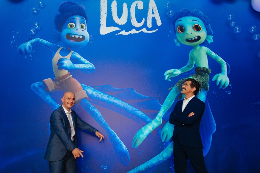 Two men stand in front of a sign advertising the movie Luca.