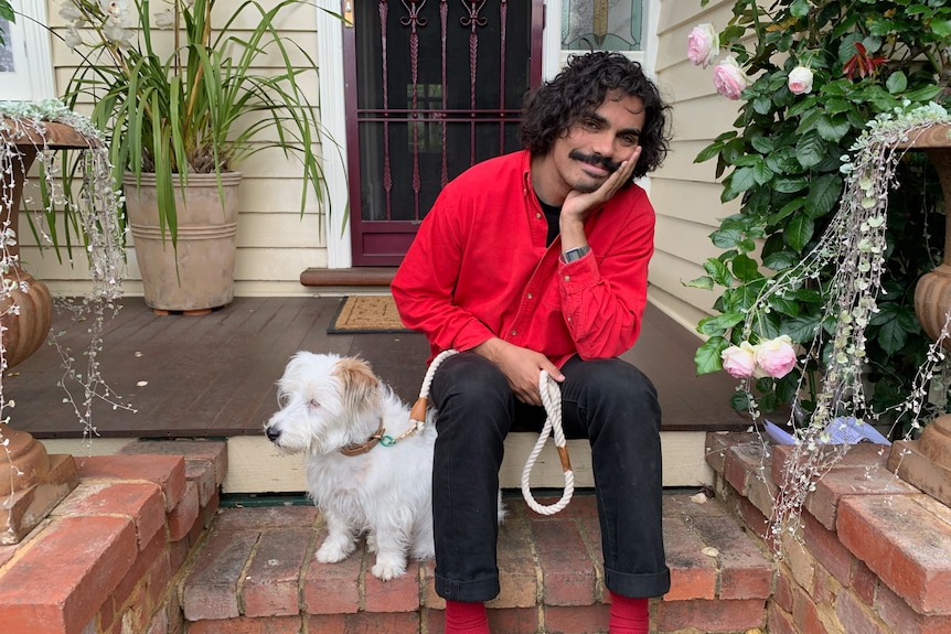 Man sitting on front step of a house with small white dog on a lead.