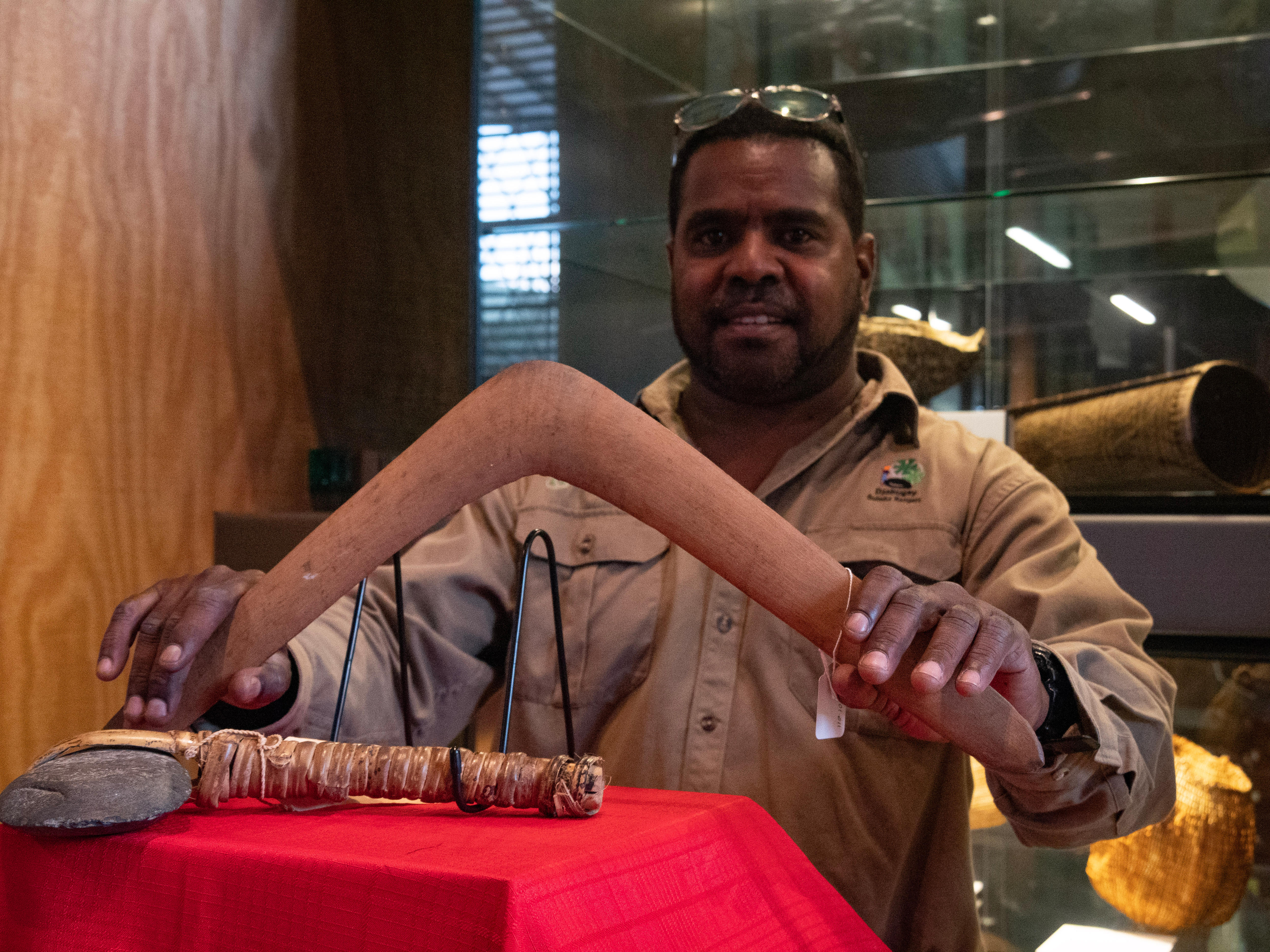 A man delicately holds a wooden boomerang, in front of it is a stone axe