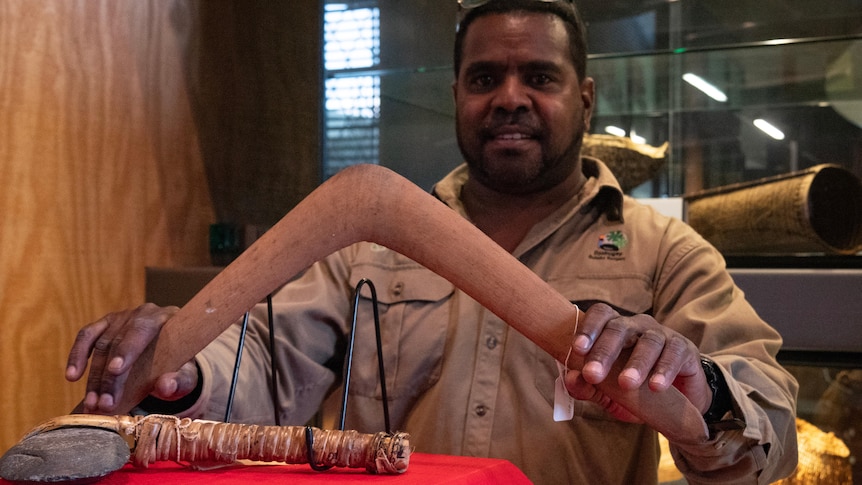 A man delicately holds a wooden boomerang, in front of it is a stone axe