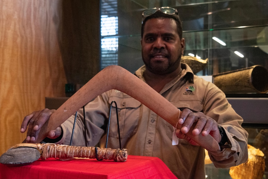 A man delicately holds a wooden boomerang, in front of it is a stone axe