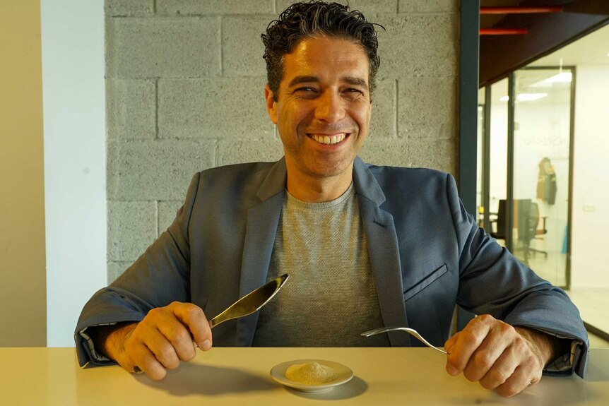 A man smiling while holding a knife and fork over a small bowl of yellow powder