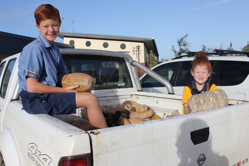 Two kids sit in the tray of a ute filled with pumpkins holding pumpkins