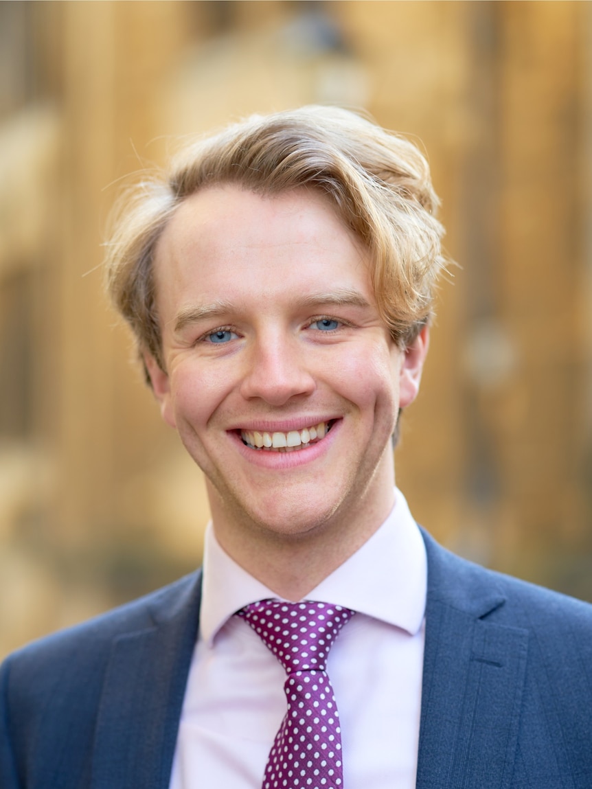 Dr Matthew Reid, with blonde hair, is pictured with a blue suit and purple spotted tie