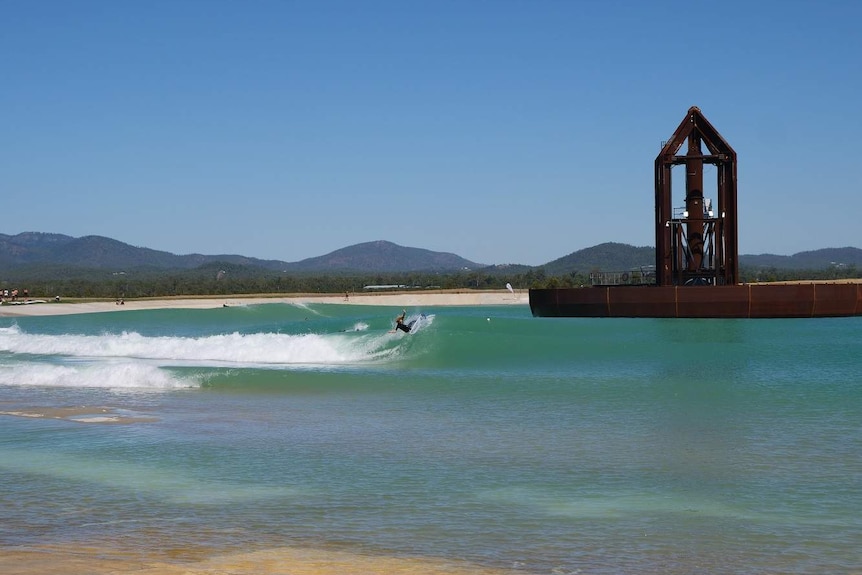 Waves crash against the bank of a wave pool while a surfer rides a wave.