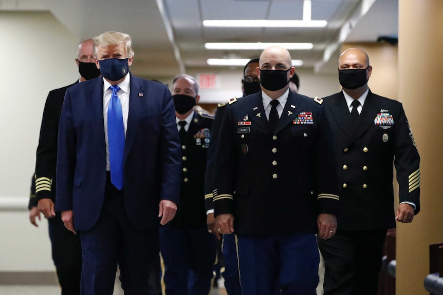 A man with blond hair wearing a suit with a blue tie walks wearing a mask down a hall with other men dressed in black suits.