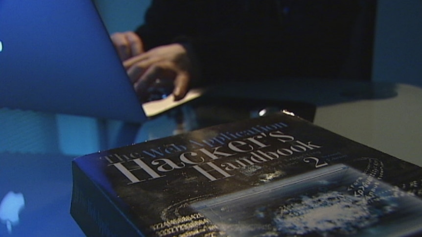 Picture of The Hacker's Handbook on a desk with an unidentified person using a laptop in the background
