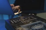 Picture of The Hacker's Handbook on a desk with an unidentified person using a laptop in the background