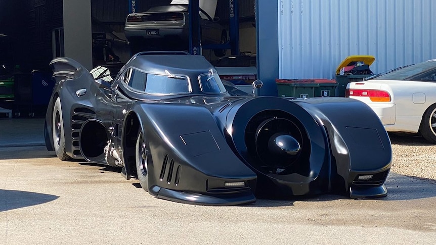 batmobile outside a workshop, rear of white car in background