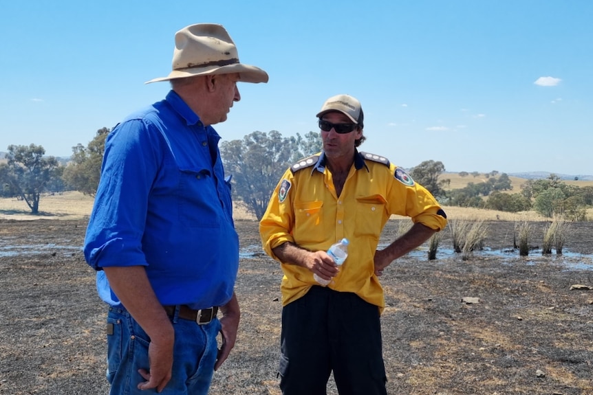 A man in a hat and blue shirt standing beside a rural firefighter. 