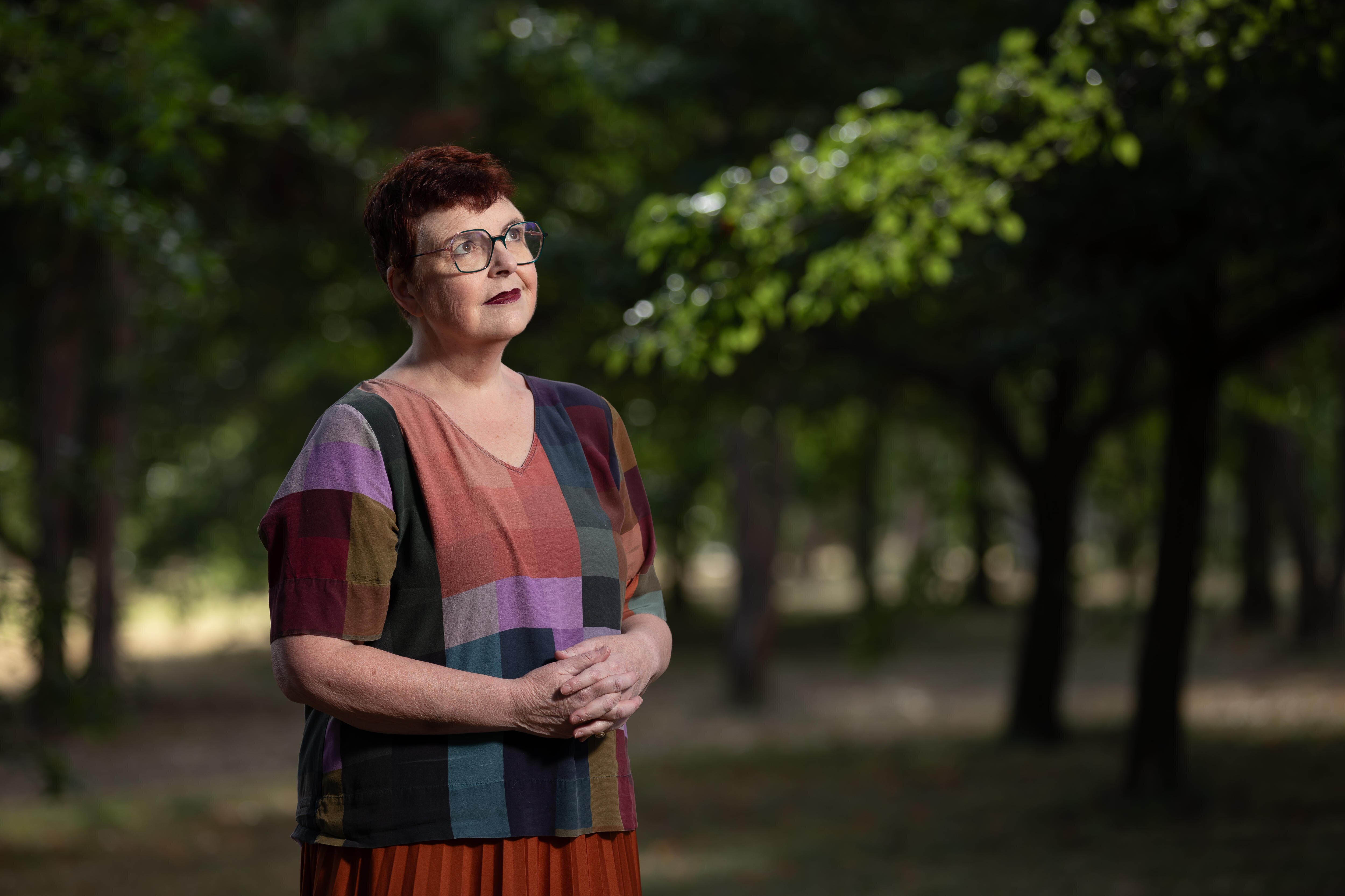 Louise wears an Autumn-coloured t-shirt and glasses and stands outside with hands clasped in front of her look towards the sky