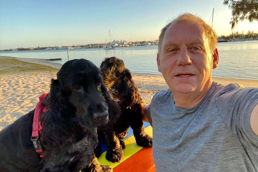 An older man takes a selfie with his two dogs on the beach.