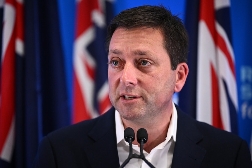 Matthew Guy stands in front of hte Australian flag and speaks into a microphone. He has a neutral expression