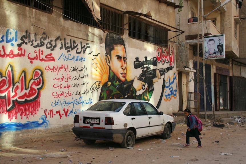 A wide photo of a young girl walking past a damaged car parked on a dirt road, in front of a mural of a young man holding a gun