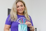 A woman with long blond hair wearing a purple shirt holding a blue booklet with a hand print on the front