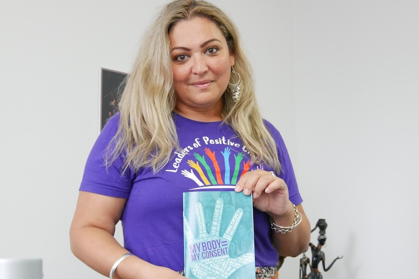 A woman with long blond hair wearing a purple shirt holding a blue booklet with a hand print on the front