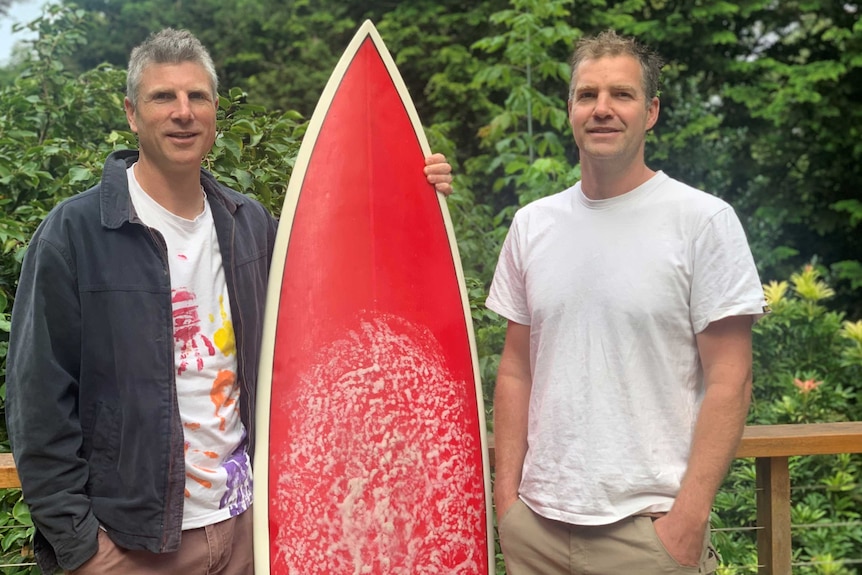 Two men in their 40s  with a surfboard.