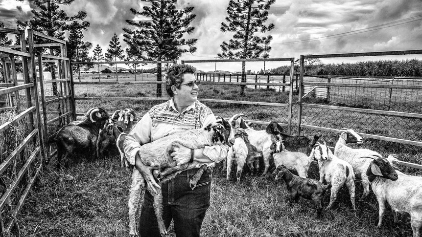 A woman holds a goat in her arms while surrounded by other goats inside their pen.