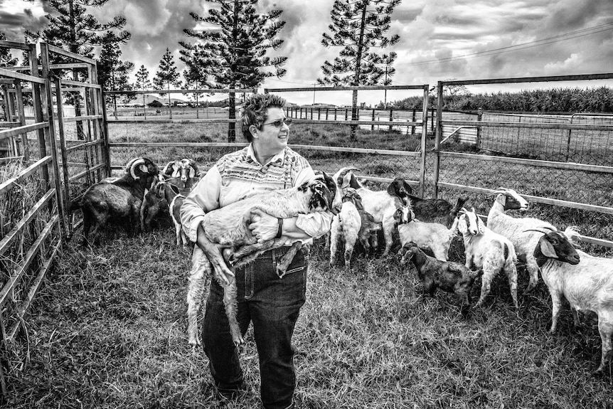 A woman holds a goat in her arms while surrounded by other goats inside their pen.