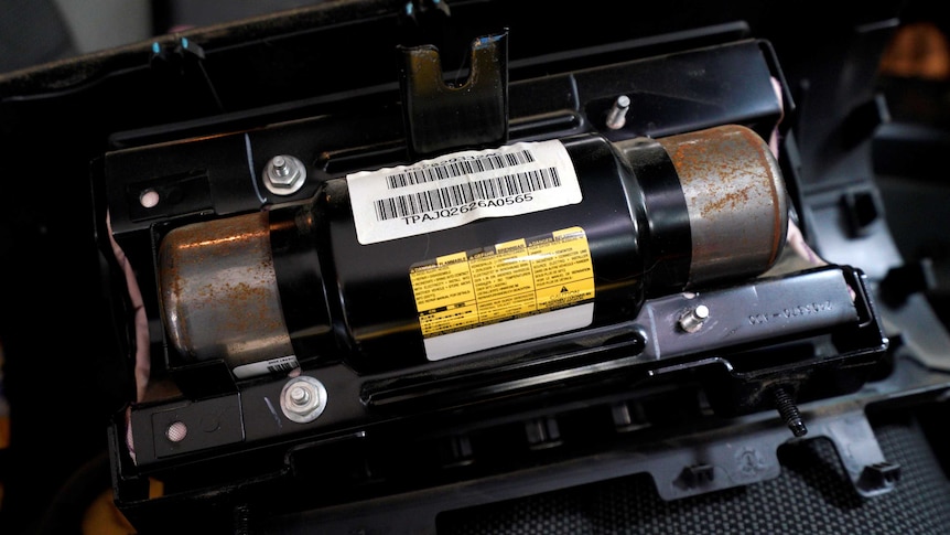 A cylindrical metal object with a barcode label and some visible surface rust sits in the engine bay of a car