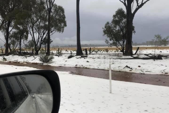 Hail has whitewashed the sides of a road and nearby paddocks, while in the background dark clouds loom