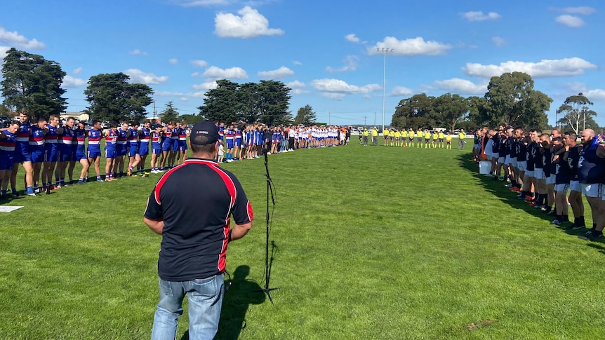 A man, seen from behind, walks to a microphone as two football teams line up.