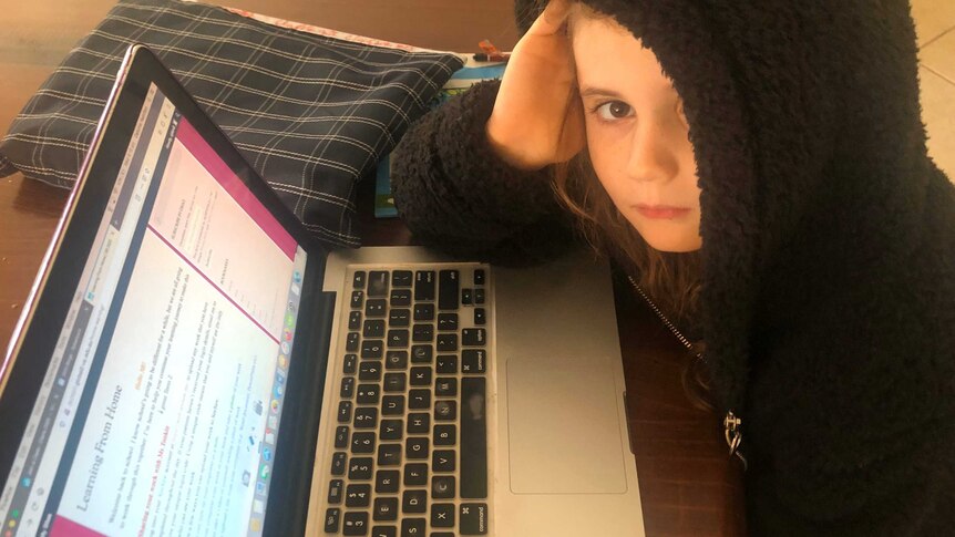 A young girl looks up at the camera as she studies at a laptop.