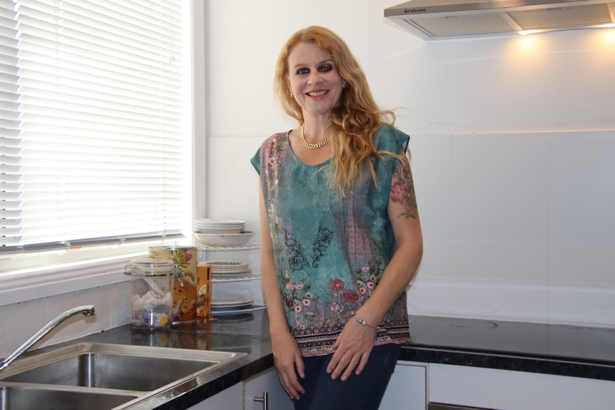 A woman with long red hair and a colourful top smiling in a kitchen