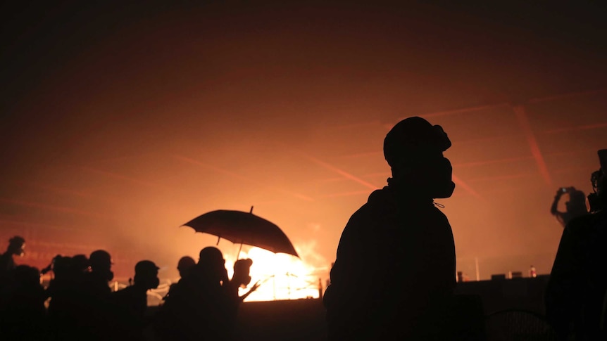 The silhouettes of police and protesters, including one with an umbrella, against an orange sky with fire in the background