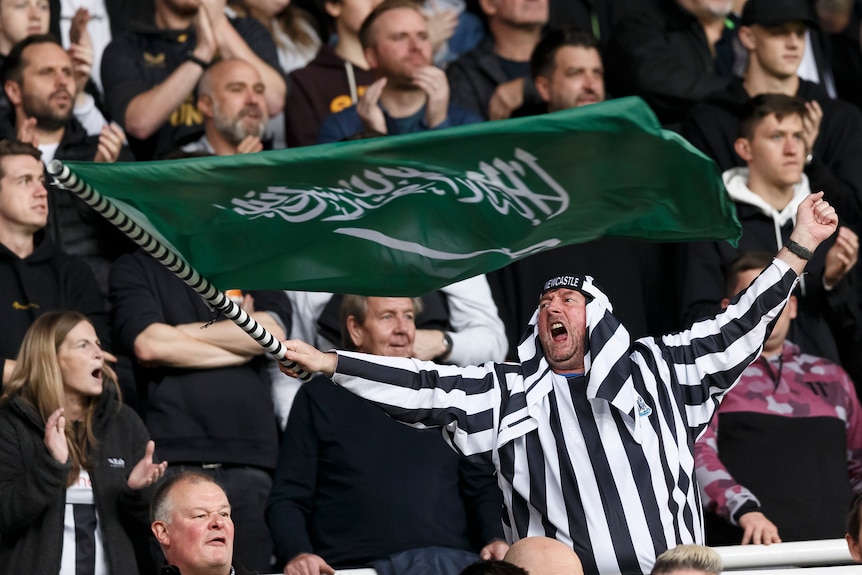 A Newcastle fan in the stands with a black and white robe and headdress waves the flag of Saudi Arabia