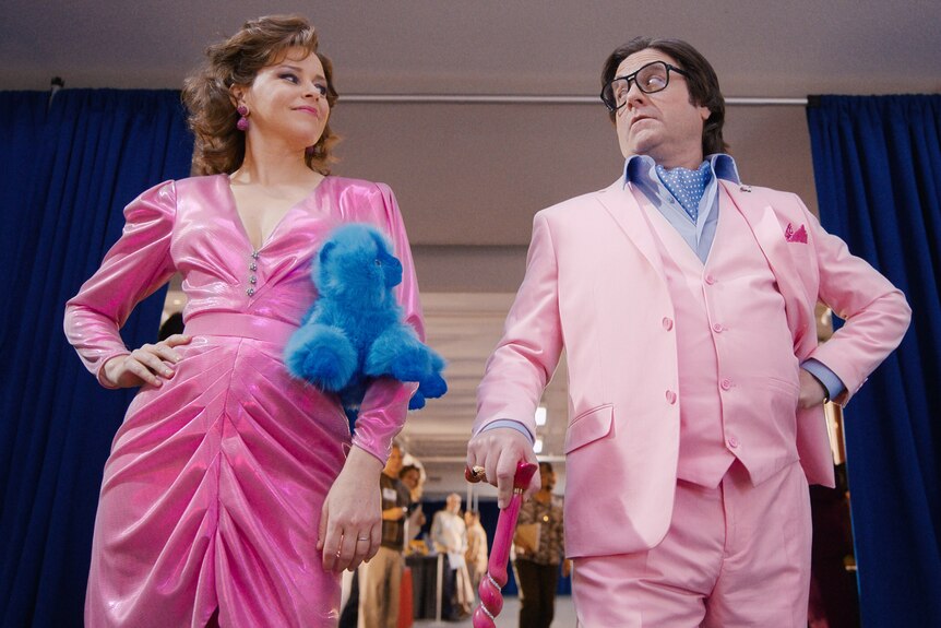 Elizabeth Banks and Zach Galifianakis stand in character, both wearing 90s clothes in bright pink, holding a blue stuffed cat