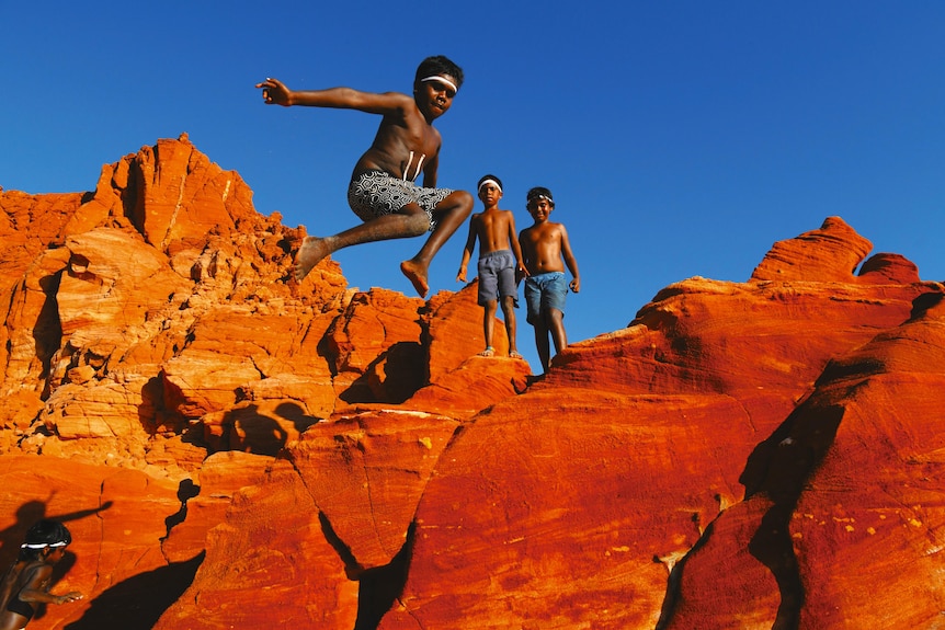 With a bright blur sky above them, young Indigenous boys wearing shorts jump across red rocks.
