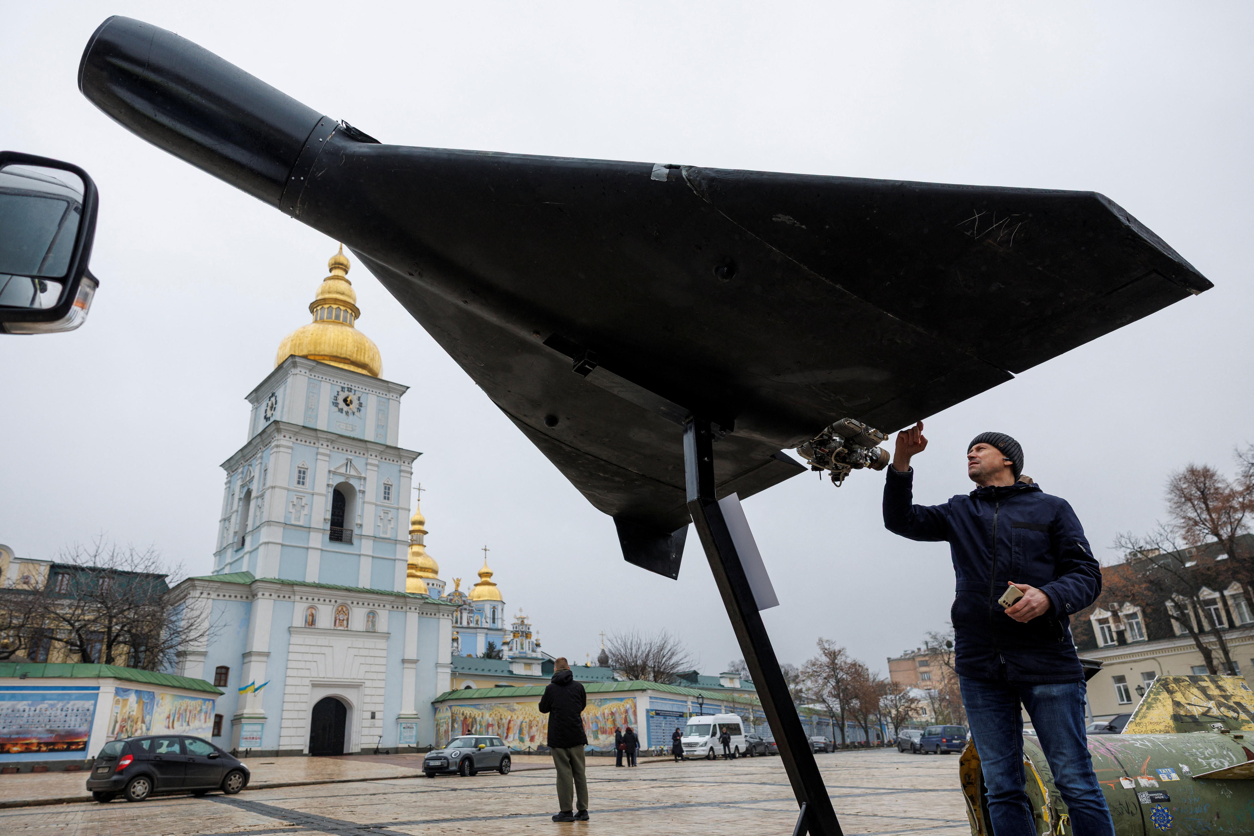 A man touches the wing of a black attack drone on display outside a Cathedral.
