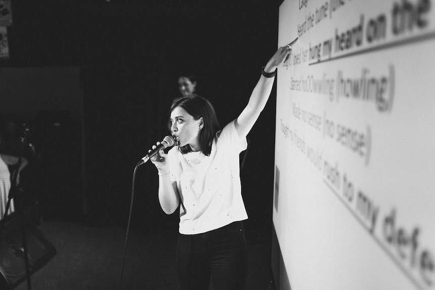 Woman stands against a projector screen which has the lyrics to a song.