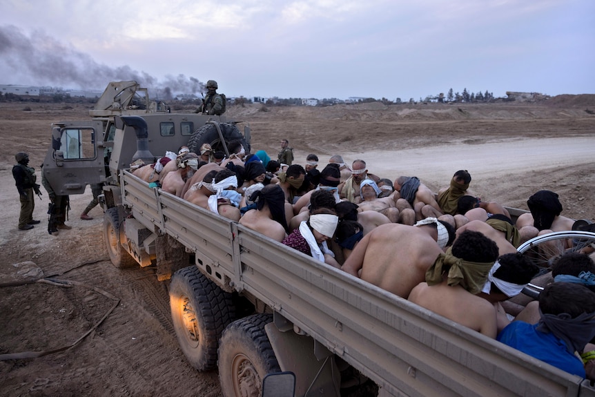 Shirtless, blindfolded men are packed into the back of a truck.