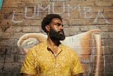 Heritier Lumumba stands in front of a wall with a mural painted on it of Patrice Lumumba.