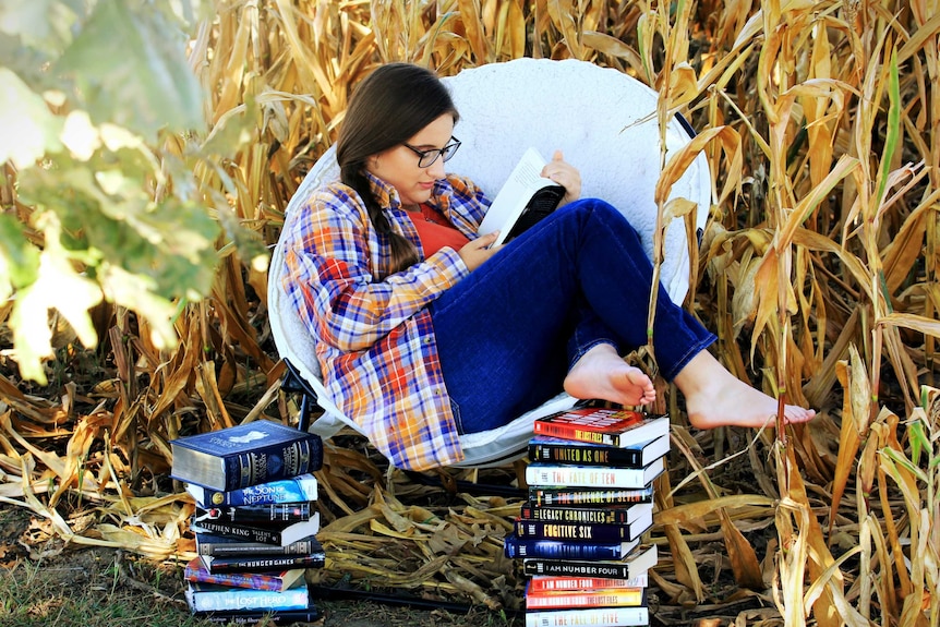 A woman with dark hair and wearing glasses sitting and reading in a field, to depict how fictional stories can change lives.