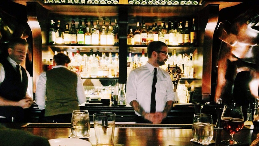 A moody bar with a waiter standing behind and bottles in the background