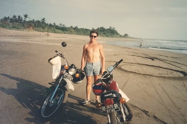 Paul Smith in his 20s smiling on a beach with motorbikes