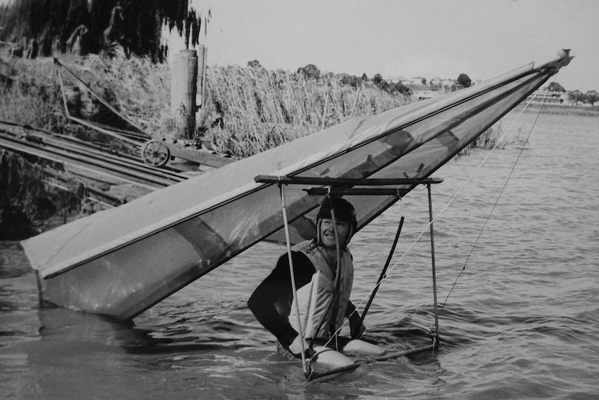 A black and white photo of a man sitting in water under an old hang glider.