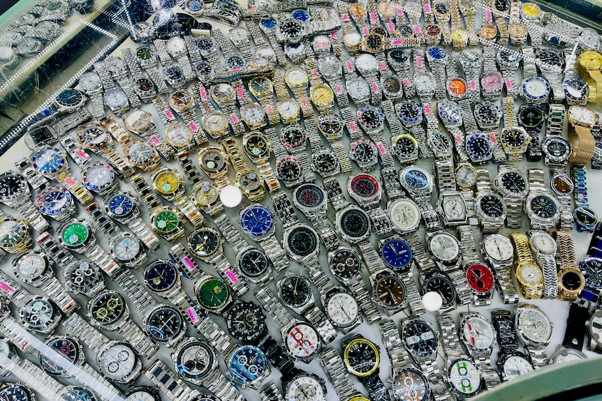 A display case shows tens of watches laid out flat, mostly gold or silver bands with different coloured faces