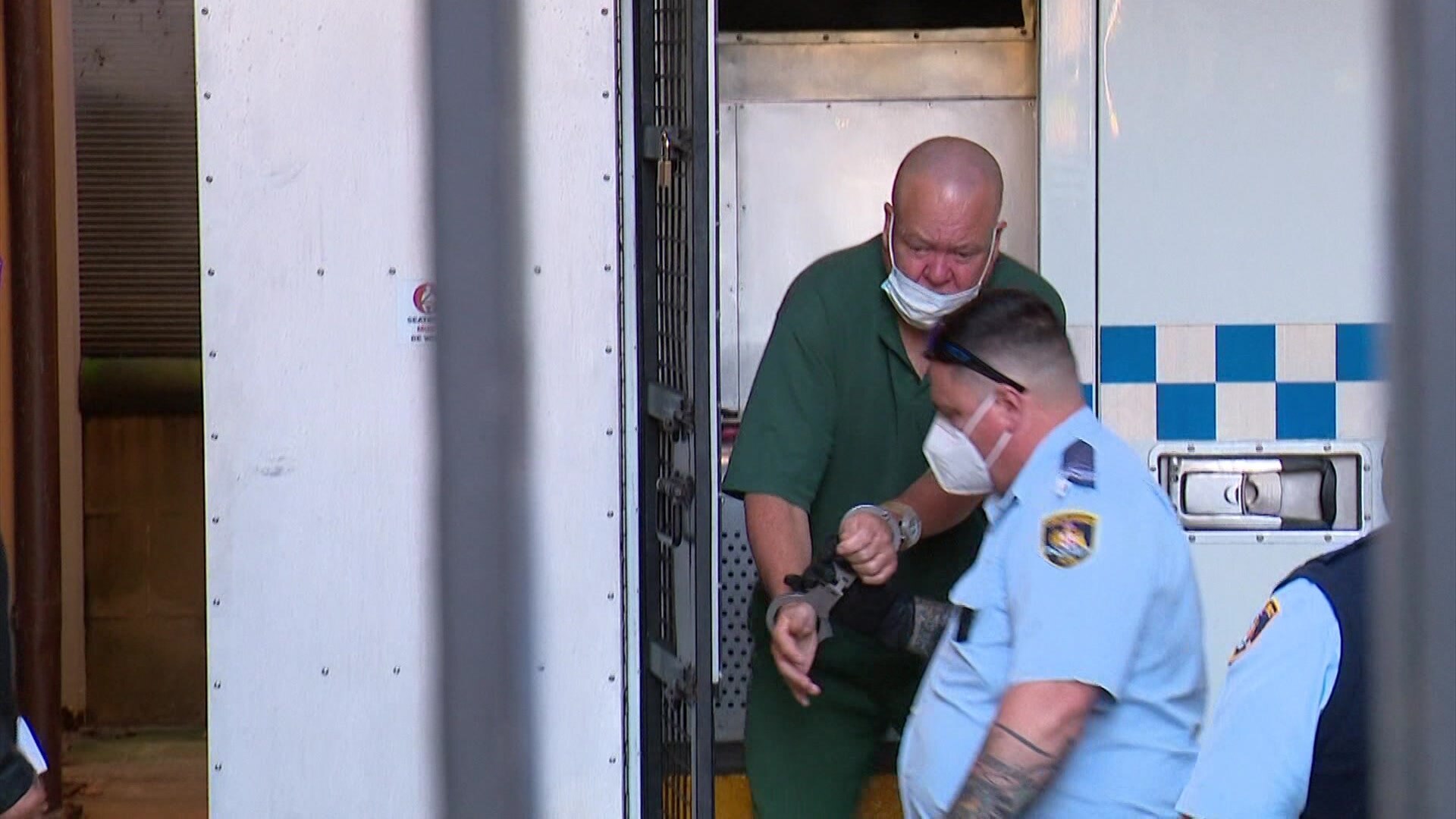 A bald man wearing handcuffs is helped from a police van
