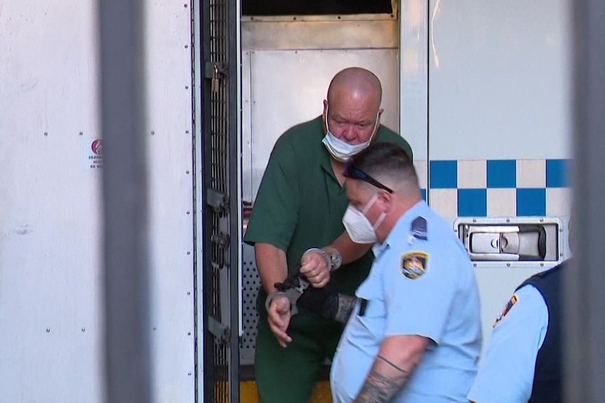 A bald man wearing handcuffs is helped from a police van