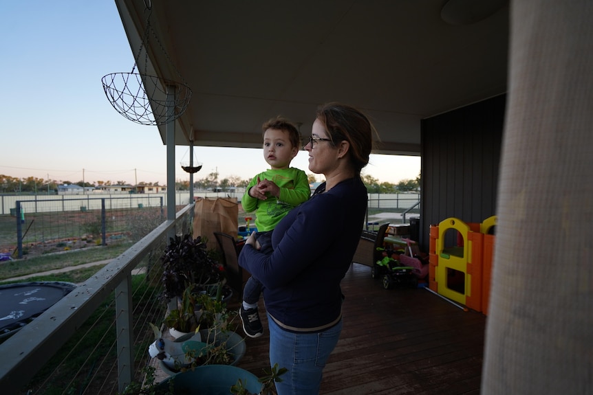 Woman stands on house deck holding young child in her arms.