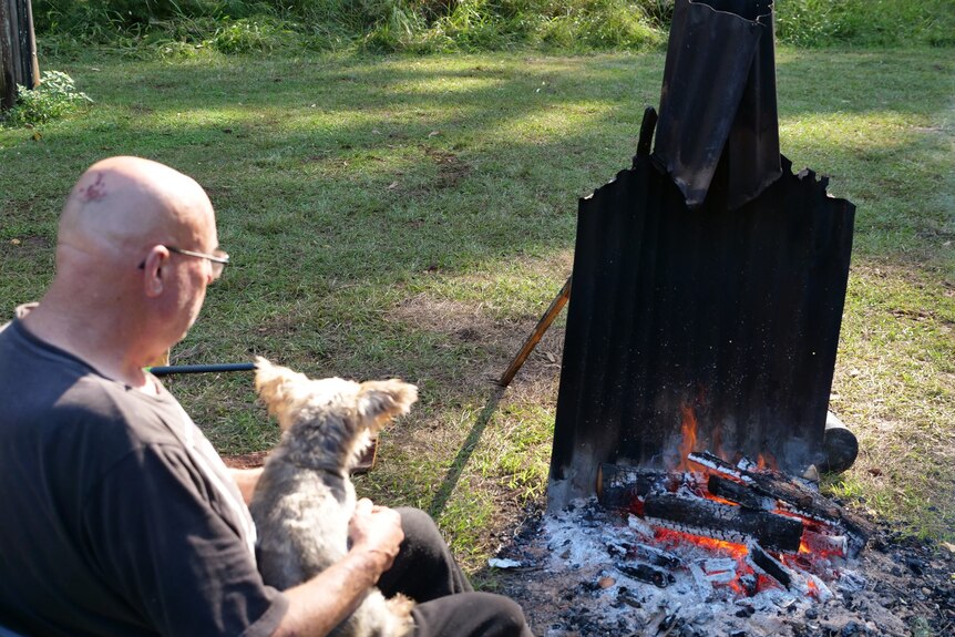 Man sitting with his dog on his lap facing the fire outside, with corrugated tin wind guard, sunlight and grassy campsite.