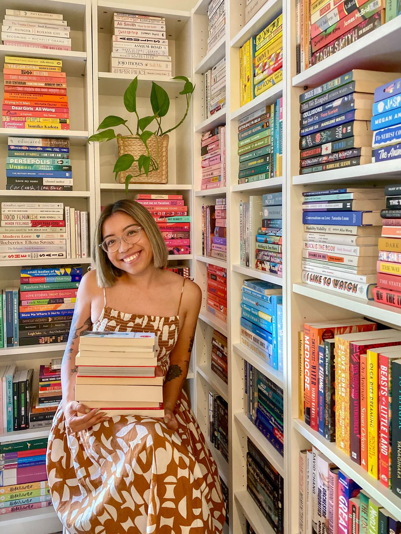 A young woman smiles as she holds a stack of books while standing in front of her towering, colourful bookshelves.