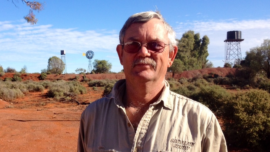 Pastoralist Steve Tonkin stands in the foreground with two tank stands and a windmill visible in the red dirt behind him